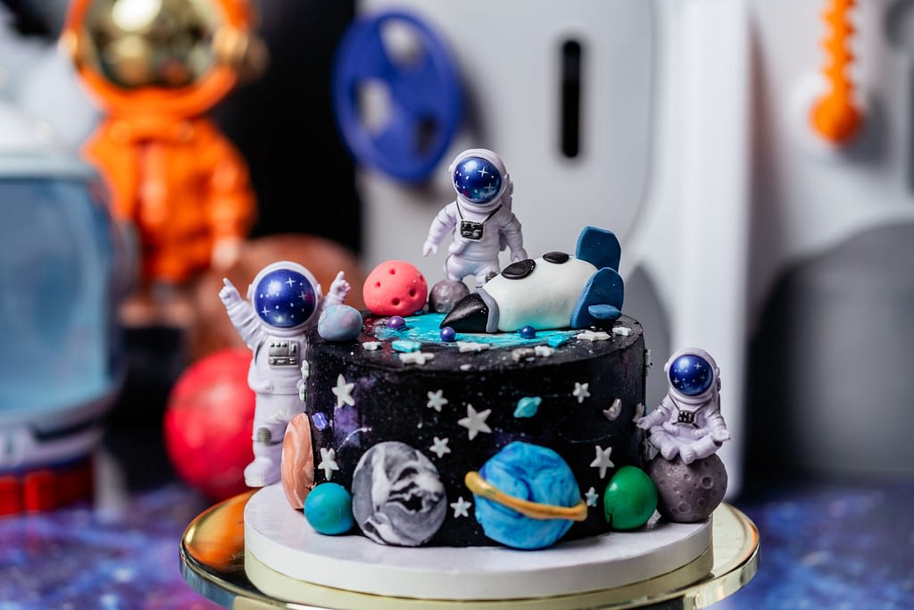 A space-themed smash cake decorated with fondant planets, stars, and astronauts, created by Moonlight Cakes from Leesburg, Virginia, sits on a round gold cake stand. The cake is designed to resemble a galaxy, with a rocket ship topper and playful astronaut figures placed around the cake. The background features space-themed decorations, enhancing the cosmic vibe of the scene. The cake is perfect for a first birthday celebration.