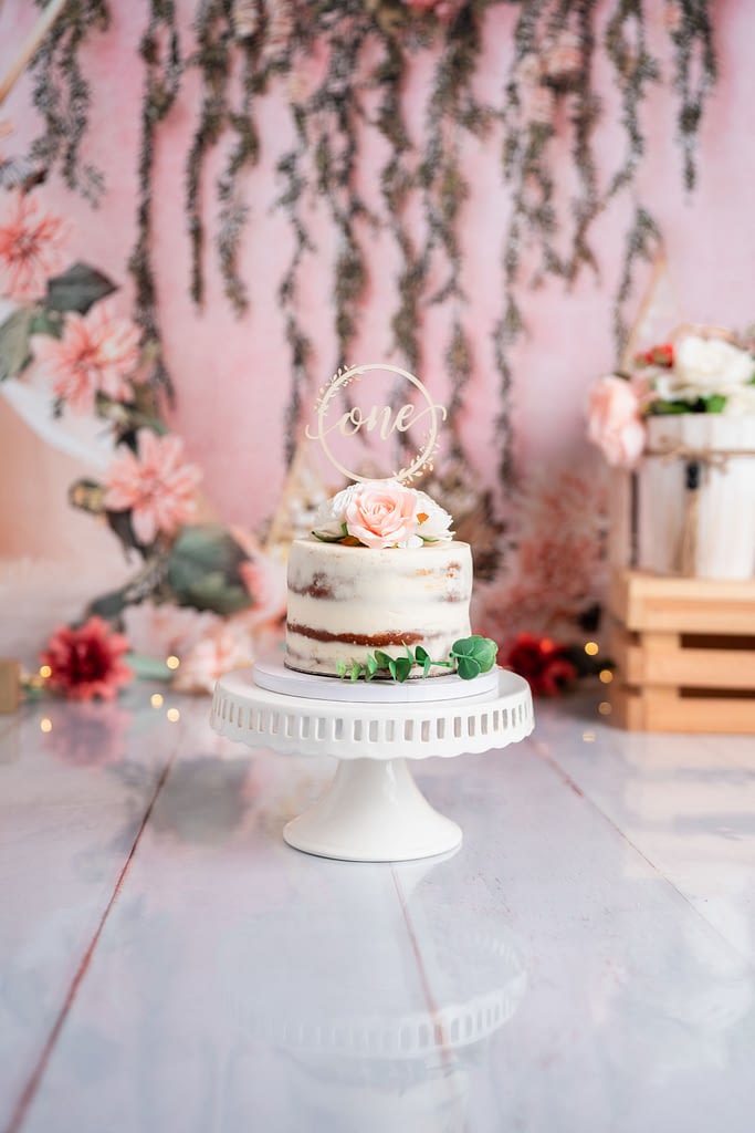 A beautifully decorated naked cake with soft white frosting sits on a white cake stand, adorned with delicate pink roses and greenery. The cake topper reads "one" in a gold, elegant script. The background features a floral arch with hanging greenery, pink flowers, and soft fairy lights, adding a whimsical and dreamy feel to the setup.