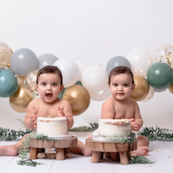 Twin boys in jumpers sitting in front of their rustic-themed cake smash setup.