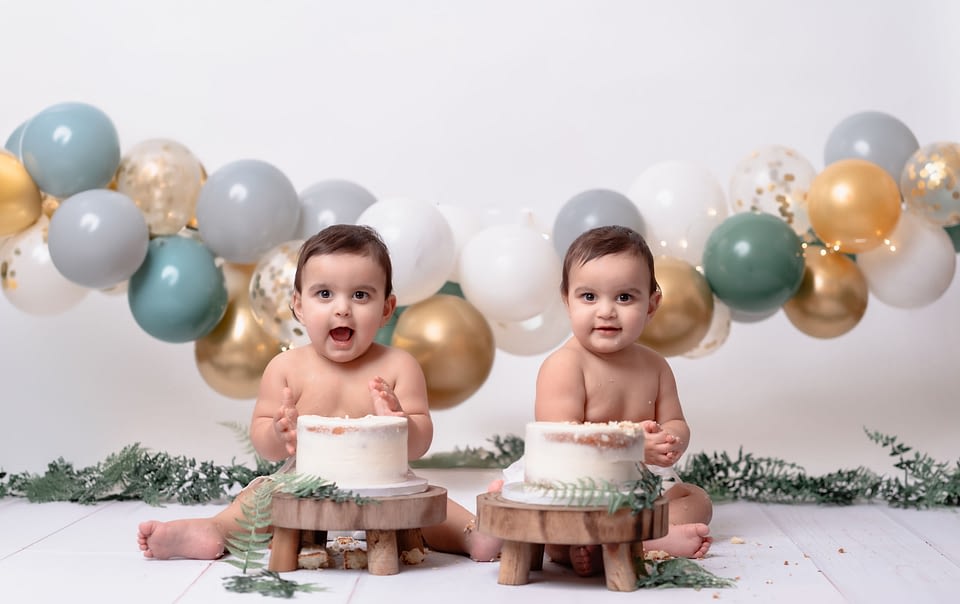 Twin boys in jumpers sitting in front of their rustic-themed cake smash setup.