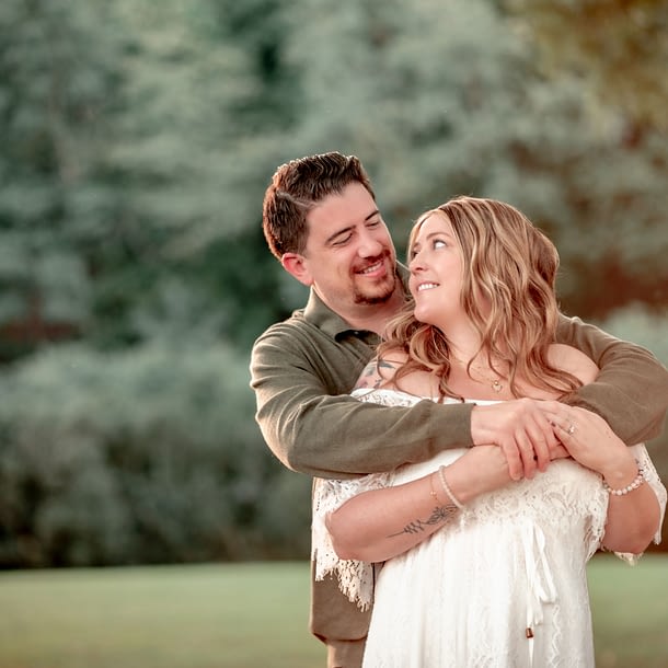 Jack embracing Cammie from behind as they both smile at each other, standing in a lush green setting at Morven Park.