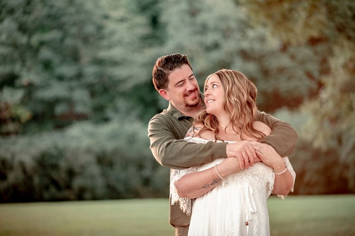 Jack embracing Cammie from behind as they both smile at each other, standing in a lush green setting at Morven Park.