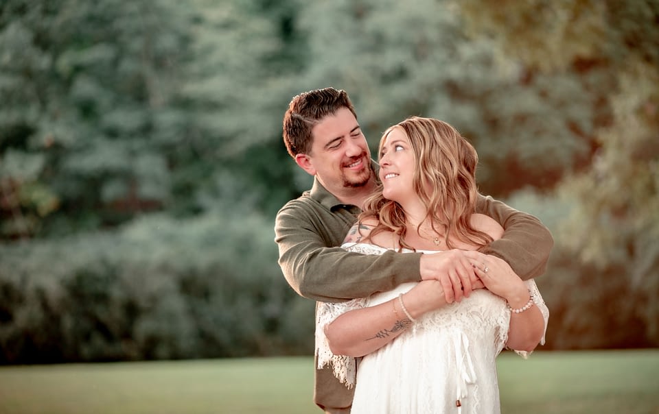 Jack embracing Cammie from behind as they both smile at each other, standing in a lush green setting at Morven Park.