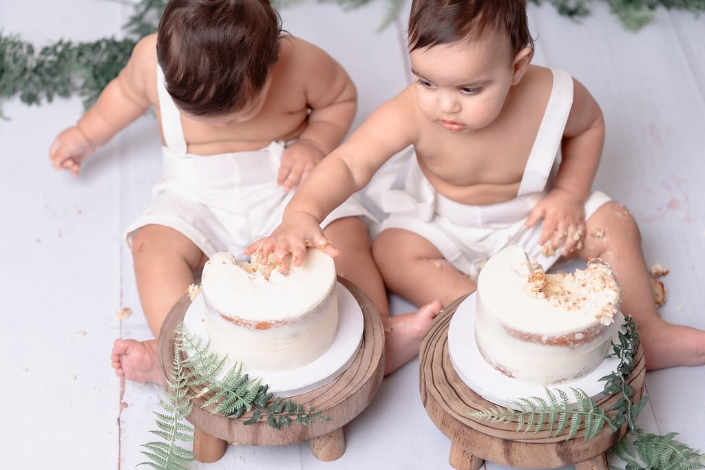 Two toddlers sitting side by side, dressed in matching white outfits, engage in a cake smash. Each child has their own small white naked cake with a minimalist design on wooden stands, with one toddler gently placing their hand into the cake. The background features subtle greenery accents and a light backdrop, creating a clean and natural aesthetic.