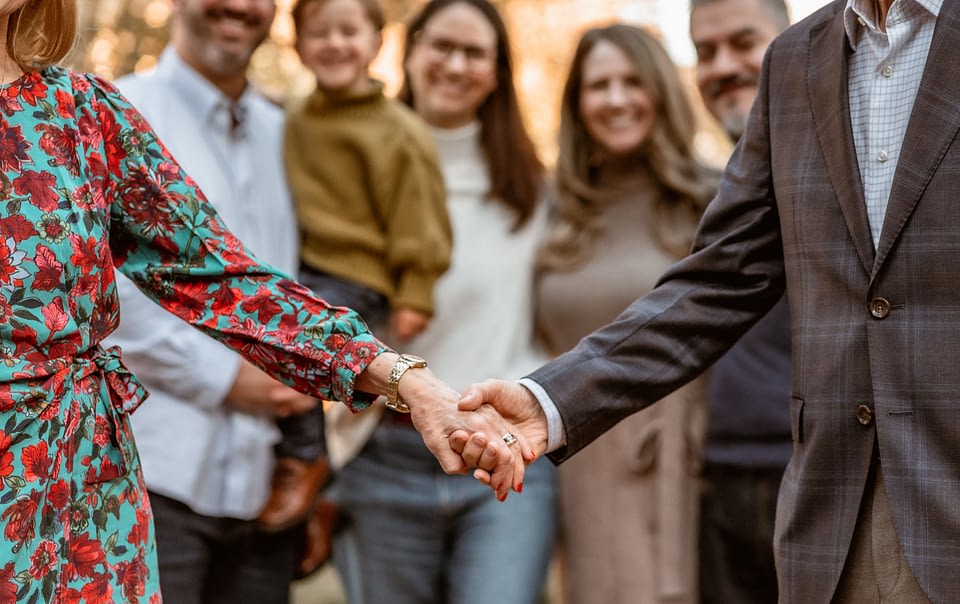 Close-up of an elderly couple holding hands during a family photo session at Carlyle House, with their smiling family softly blurred in the background.