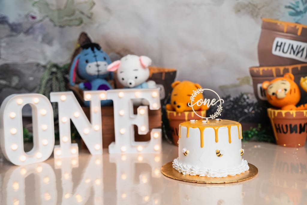 A honey-themed cake with golden drip icing and tiny bee decorations sits on a gold cake board, topped with a circular "one" cake topper. The background features plush characters from Winnie the Pooh, including Eeyore and Piglet, with a rustic backdrop of "hunny" pots and a marquee "ONE" sign lit up in white lights.