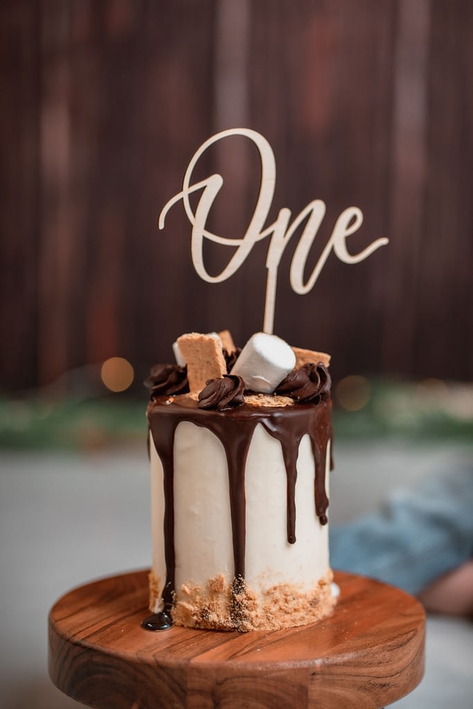 A beautifully decorated smash cake photographed by Kristina Yazmin Photography, featuring a white cake with rich chocolate drip frosting. The cake is topped with graham crackers, marshmallows, and chocolate swirls, evoking a s'mores theme. A wooden topper spelling "One" is placed on top, celebrating a first birthday. The cake is displayed on a wooden cake stand with a blurred, warm-toned background, perfect for a cake smash session in Kristina's Ashburn studio.